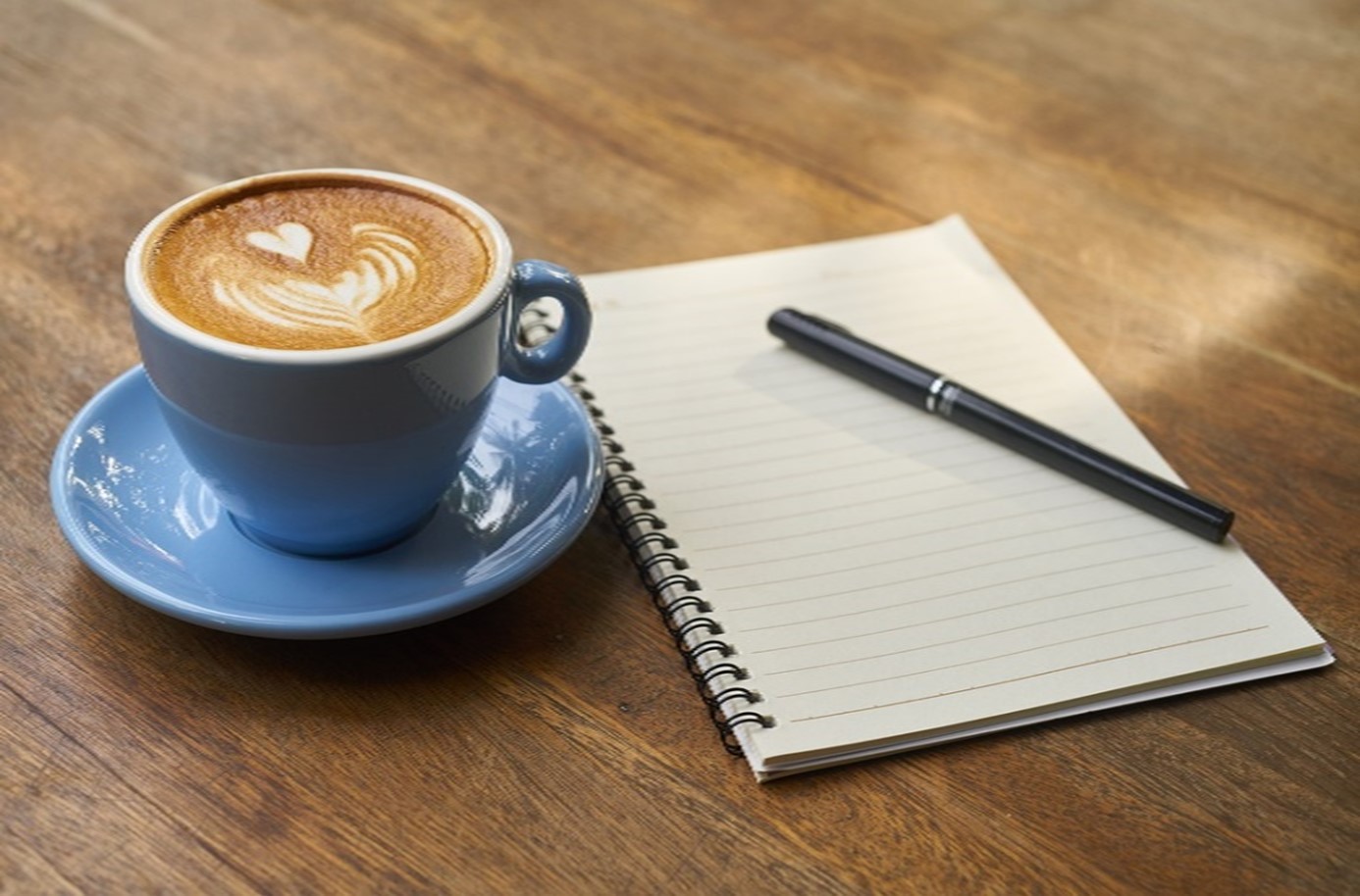 Eine Tasse Cappuccino auf einem Tisch, daneben Schreibblock und Kugelschreiber.