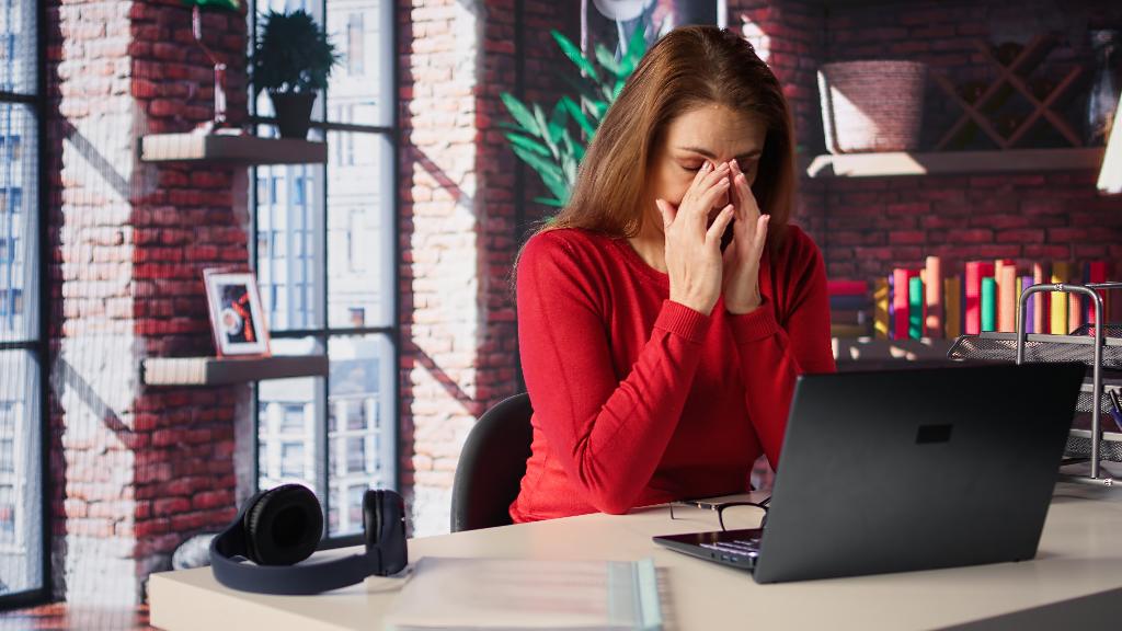 Eine Frau sitzt am Fenster in ihrem Loft am Laptop und reibt sich müde ihre Augen.