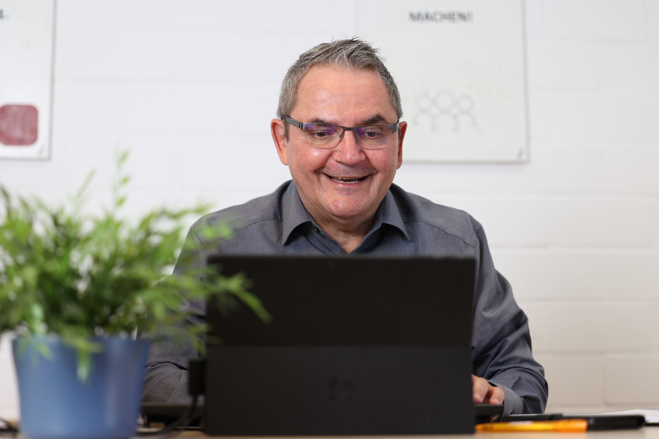 Mann arbeitet lachend am Laptop, Pflanze im Vordergrund.