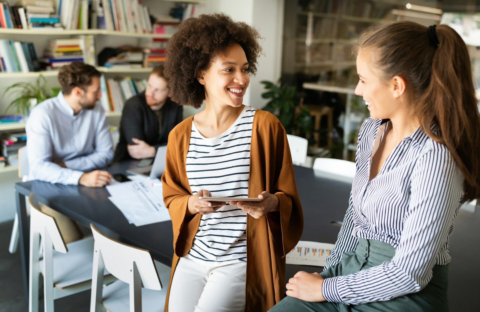 Lächelnde Frauen im Büro, Männer arbeiten im Hintergrund.